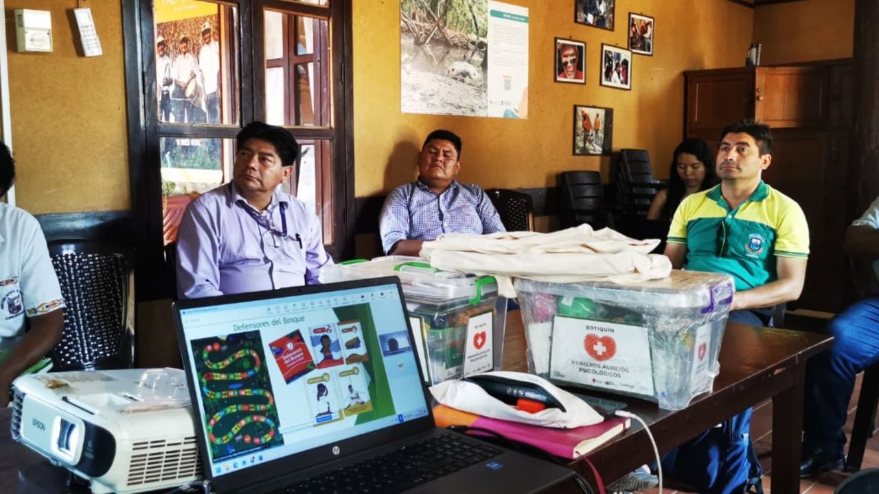 A group of people seated around a table with a laptop and projectors, discussing materials in a room with wooden decor.