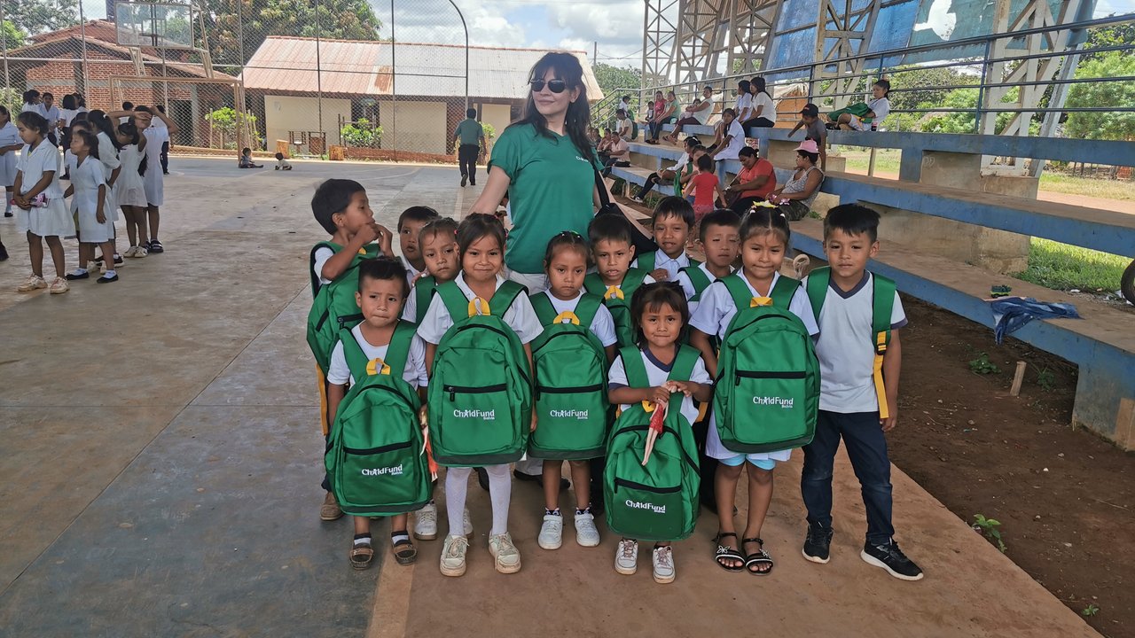 A group of children in uniforms holding green backpacks, with adults and other children in the background.