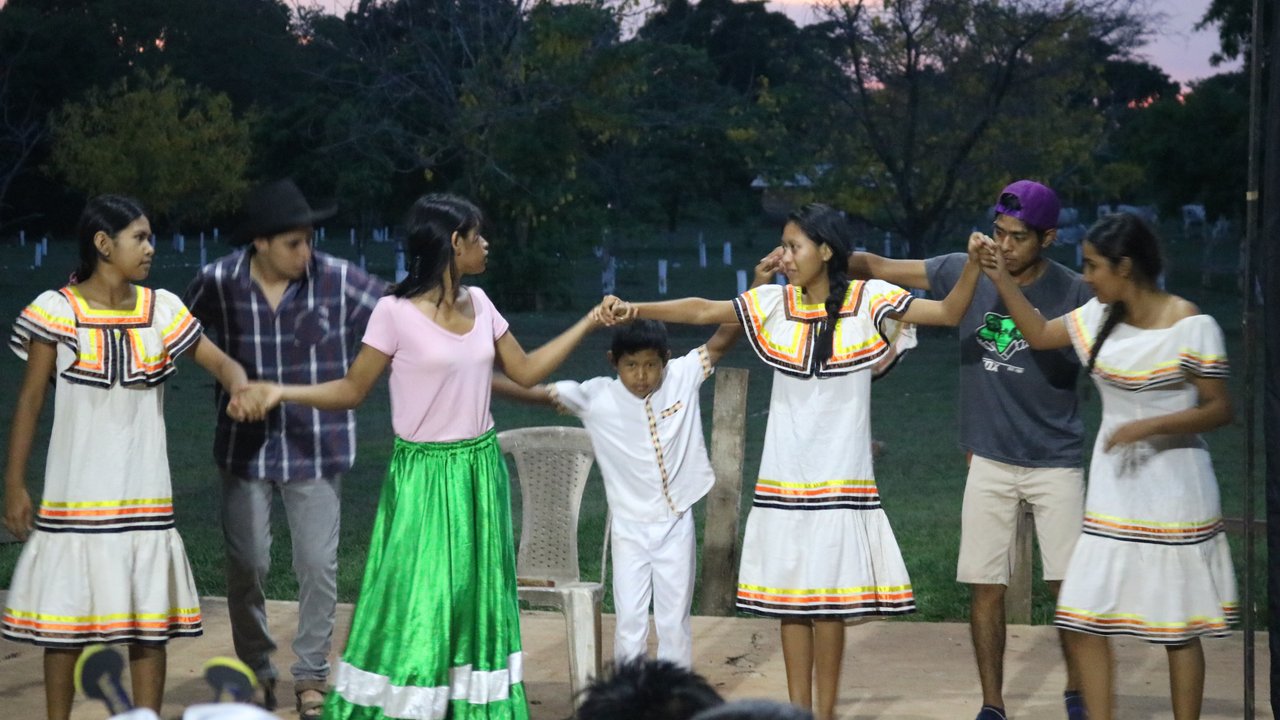 A group of children and adults perform a dance outdoors, wearing traditional clothing, with trees in the background.