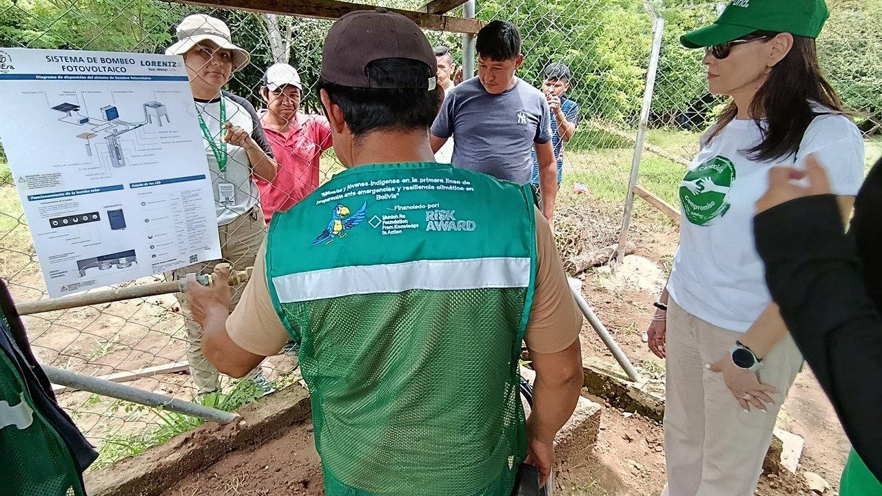 Un grupo de personas observa un cartel sobre un sistema de bombeo fotovoltaico en un entorno natural.