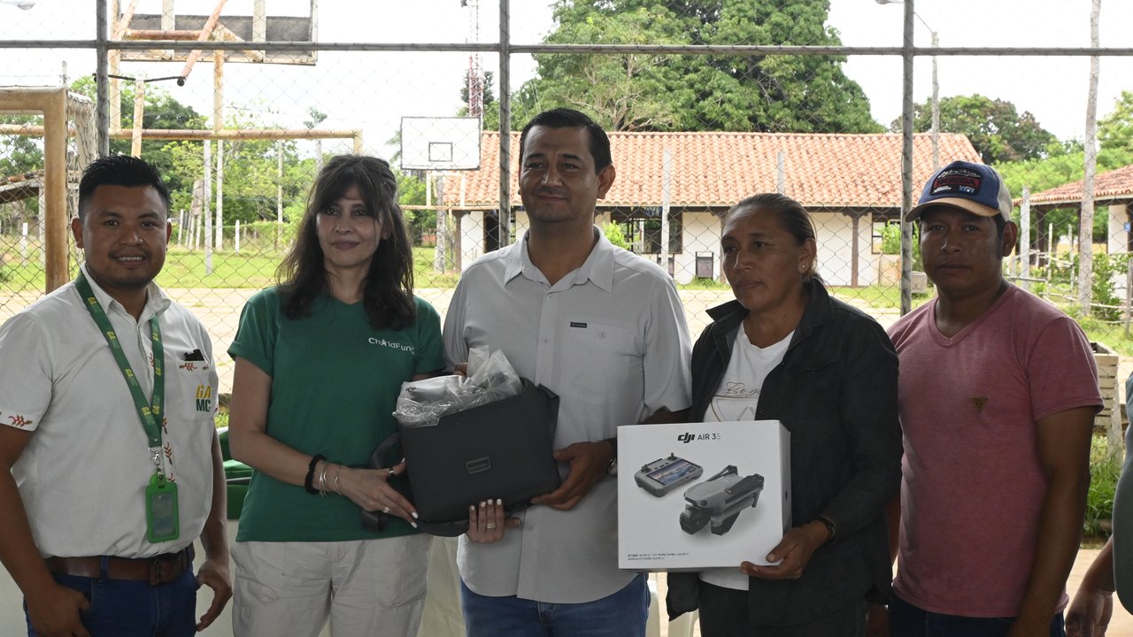 A group of five people stands together, holding a drone box and other items, with a sports court in the background.
