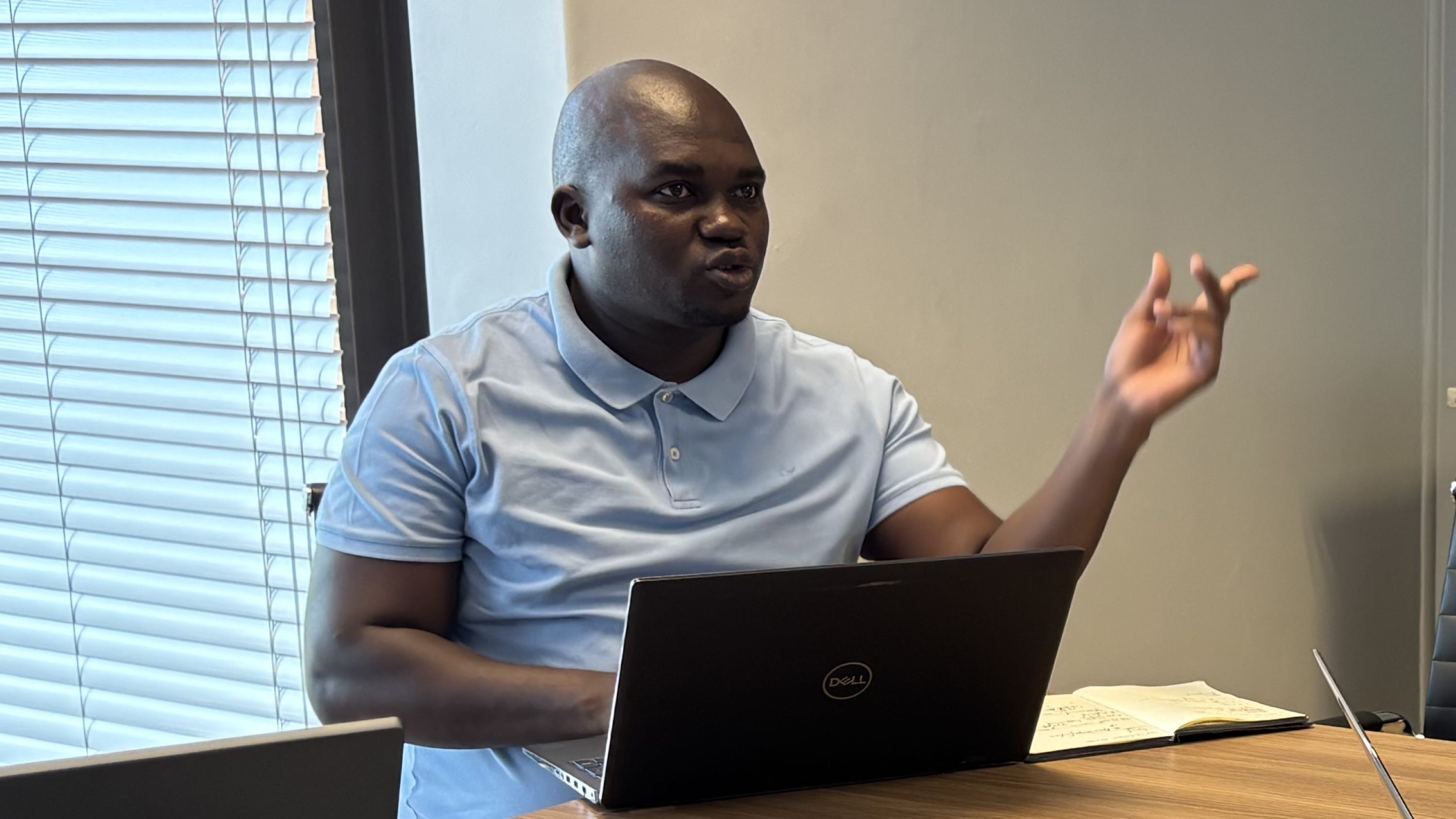 A person in a light blue shirt gestures while sitting at a table with a laptop and notebook.