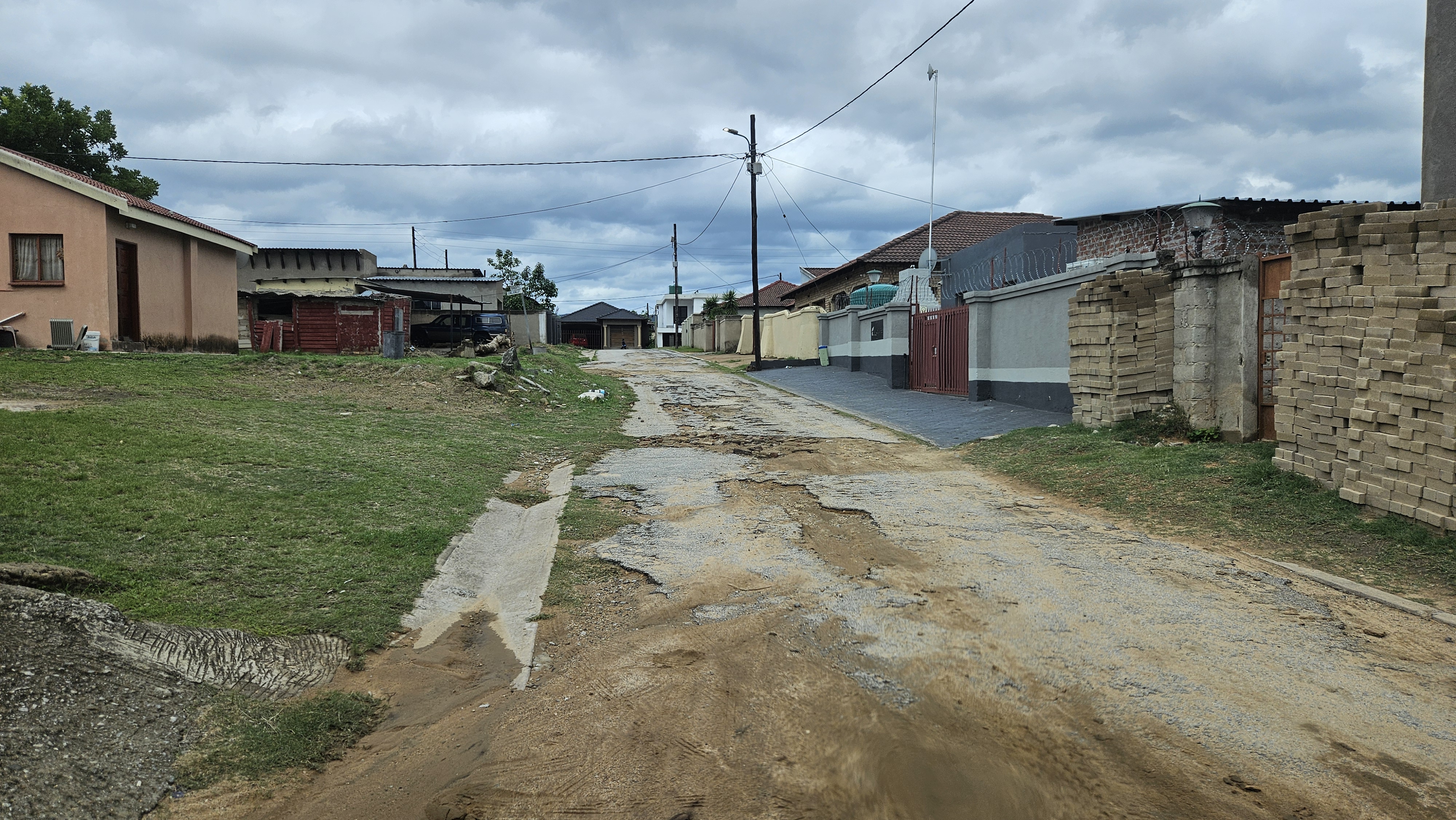 A dirt road lined with houses, some with fences, under a cloudy sky.