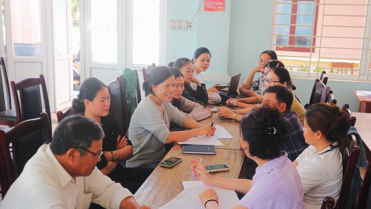 A group of women and men sit around a wooden table in a bright room, talking and taking notes. Some have notebooks and phones in front of them, while one person uses a laptop. Everyone appears attentive and engaged in the conversation.