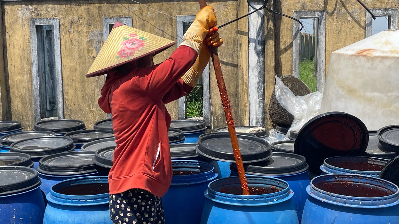 A woman wearing a traditional woven hat, a red jacket, and gloves stirs the contents of a large blue barrel with a long wooden stick. Many similar barrels are arranged around her, set against an old yellow wall with windows.