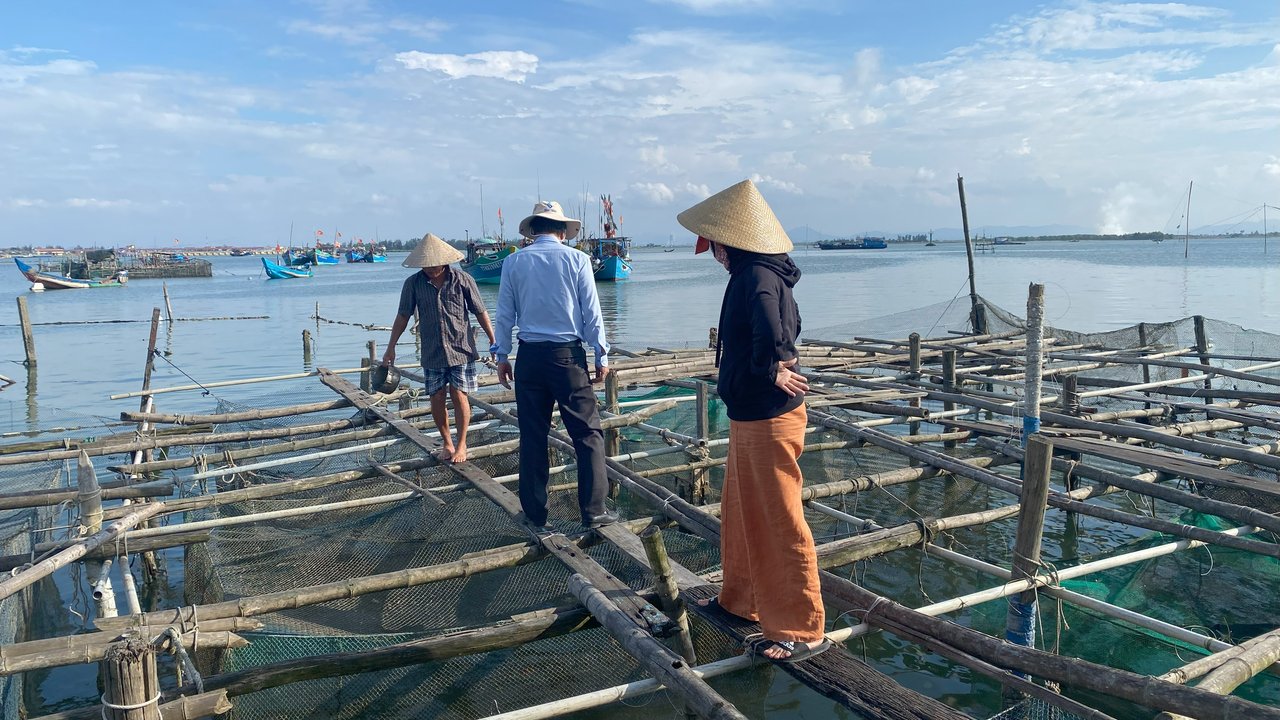 Three people stand on a wooden cage farming platform above the water in Phuong Thuan An. Two wear traditional conical hats, and fishing boats are visible in the background. The sea is calm, and the sky is partly cloudy in the late afternoon light.