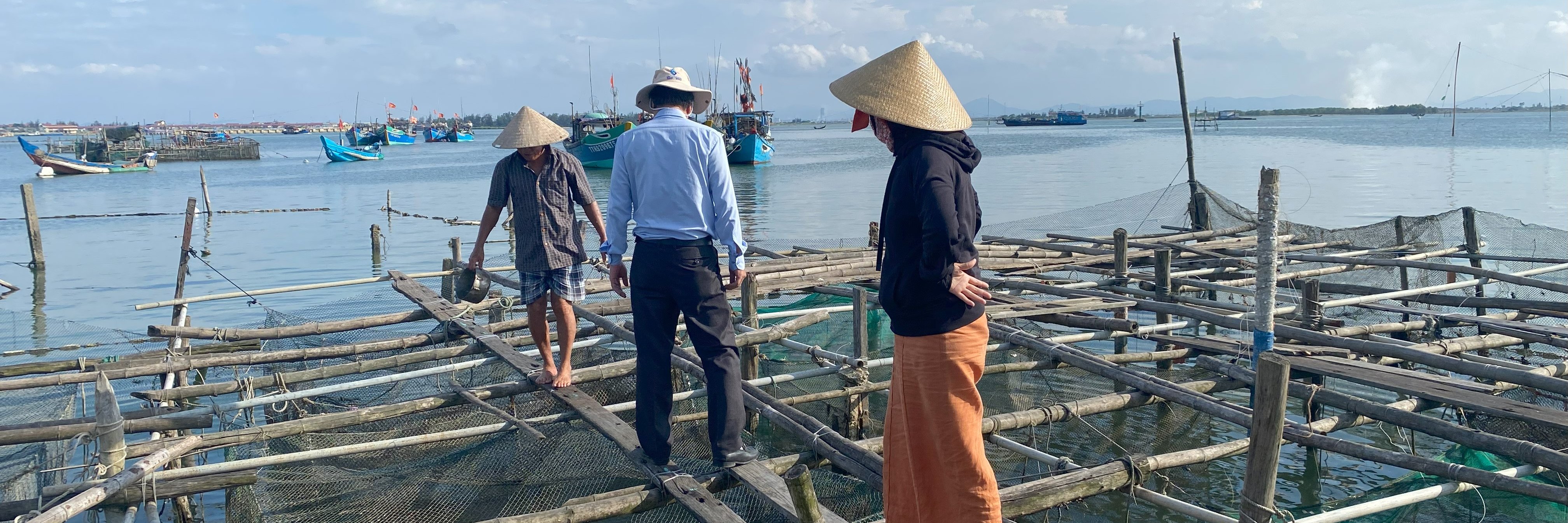Three people stand on a wooden cage farming platform above the water in Phuong Thuan An. Two wear traditional conical hats, and fishing boats are visible in the background. The sea is calm, and the sky is partly cloudy in the late afternoon light.