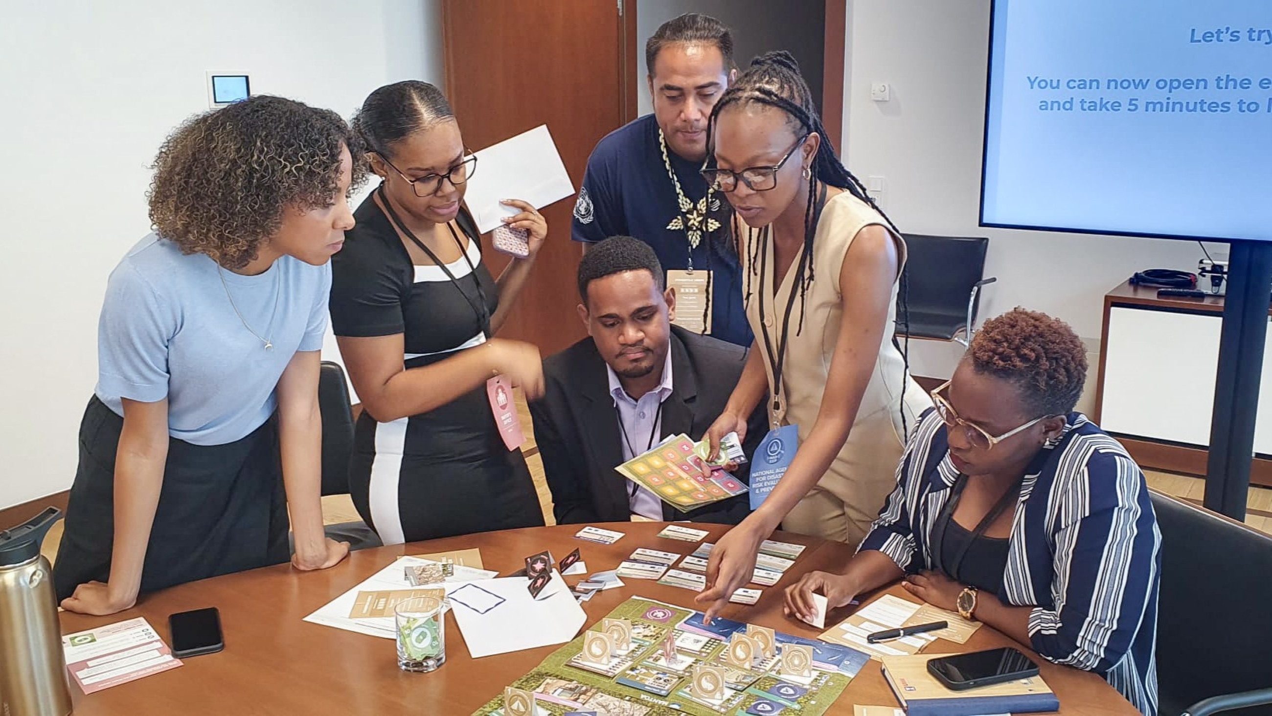 A group of six people gather around a table with game materials and a presentation screen in a meeting room.