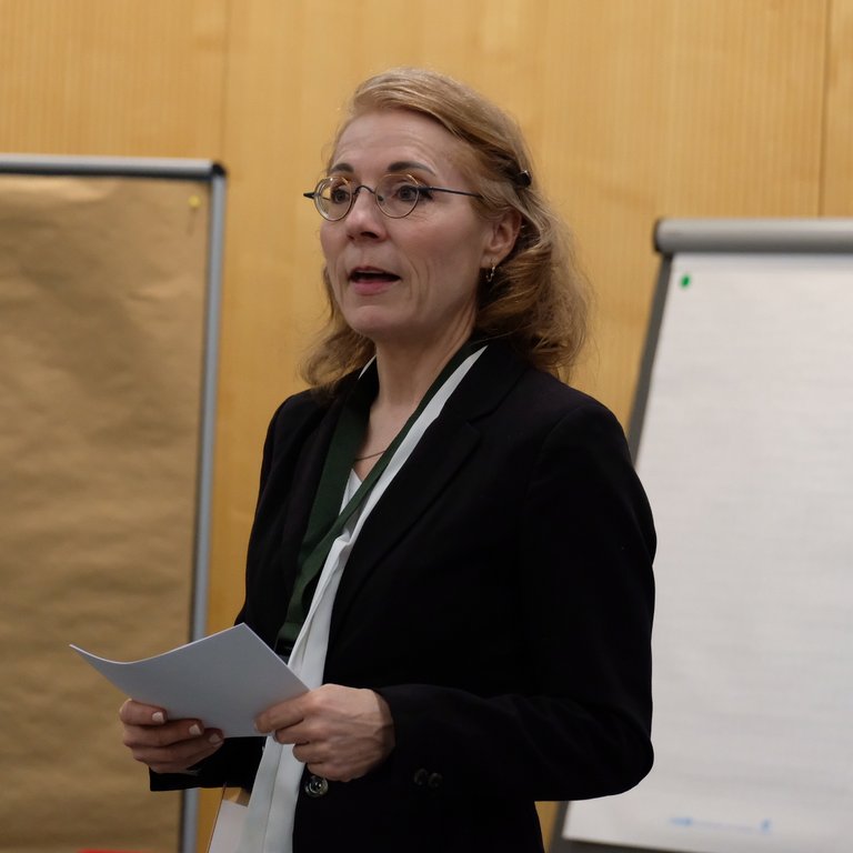 A person in a black suit holds papers, standing in front of a flip chart and a brown backdrop.