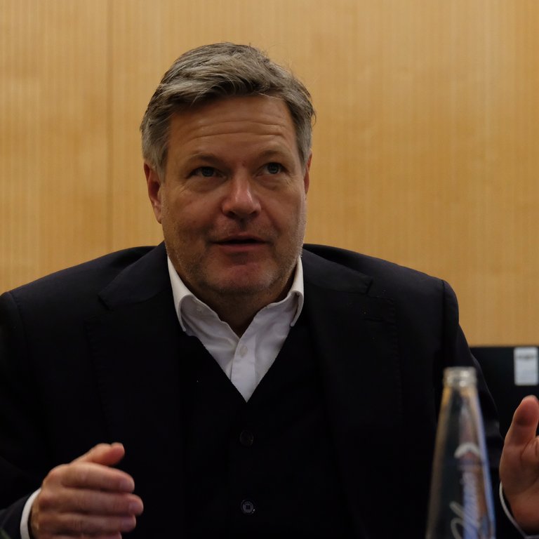A person in a suit gestures while seated at a table with a water bottle and wooden paneling in the background.